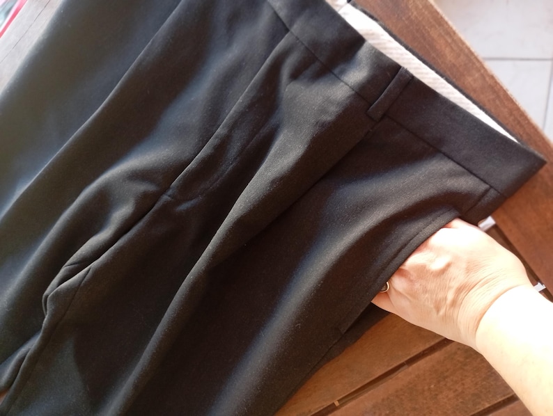 May include: A pair of black trousers featuring a structured waistband with belt loops, a front zip fly closure, and side pockets. The fabric has a smooth, matte finish, and the design includes a clean, tailored cut with visible seam detailing.