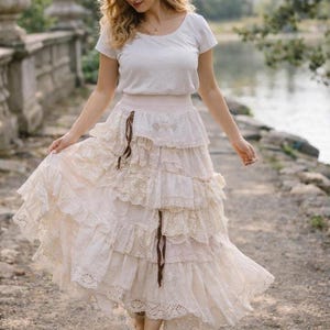 Può includere: Una gonna a balze color crema con dettagli in pizzo e occhielli, abbinata a una maglietta bianca. La gonna ha uno stile bohémien fluido, con un accento di nastro marrone. L'outfit è indossato all'aperto, suggerendo un'occasione casual o speciale.