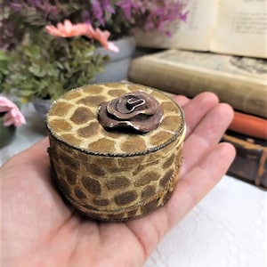 Peut inclure: Une petite boîte ronde avec un motif imprimé de girafe brun et beige. La boîte est ornée d'une bordure dorée et d'une rose brune sur le dessus.