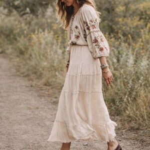 May include: A cream-coloured tiered maxi dress with floral embroidery on the sleeves and bodice. The outfit includes a brown fedora hat and brown leather loafers. The dress is flowing and appears to be made of a lightweight fabric.