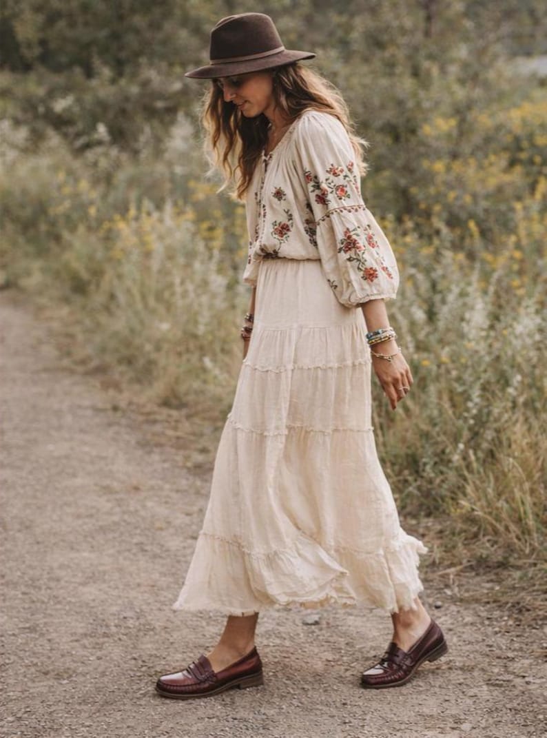 May include: A cream-coloured tiered maxi dress with floral embroidery on the sleeves and bodice. The outfit includes a brown fedora hat and brown leather loafers. The dress is flowing and appears to be made of a lightweight fabric.