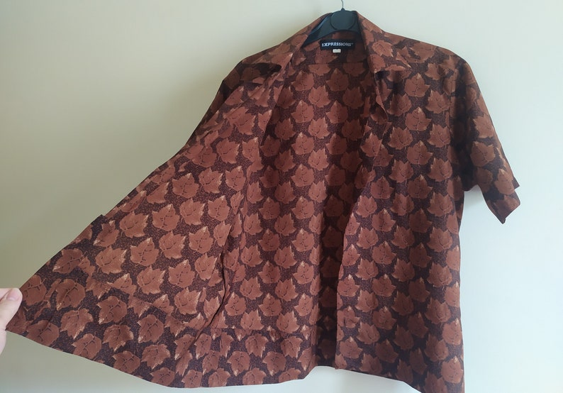Puede incluir: Una camisa marr&oacute;n de manga corta con botones y un patr&oacute;n repetitivo de peque&ntilde;os gatos marrones. La camisa tiene cuello y un bolsillo en el lado izquierdo. La etiqueta de la camisa dice "EXPRESSIONS".