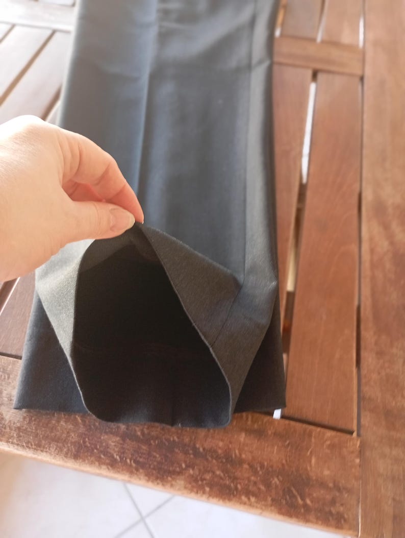 May include: A pair of dark charcoal grey trousers featuring a straight-leg cut and a clean, pressed crease down the front. The fabric has a smooth, matte texture, and the hem is finished with a simple, straight edge, showing the interior lining.