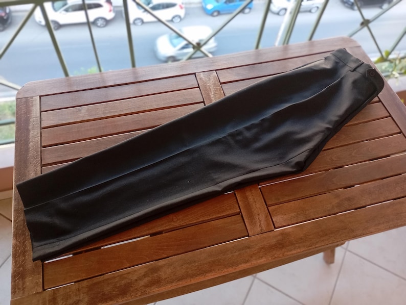 May include: A pair of solid black trousers featuring a tailored fit, a structured waistband with belt loops, and a clean, smooth finish. The trousers are folded flat, showcasing a straight leg design with a visible crease running down the centre.