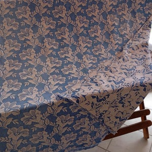 May include: A blue and white floral patterned fabric with a repeating design of stylized flowers. The fabric is folded in half and draped over a wooden chair.