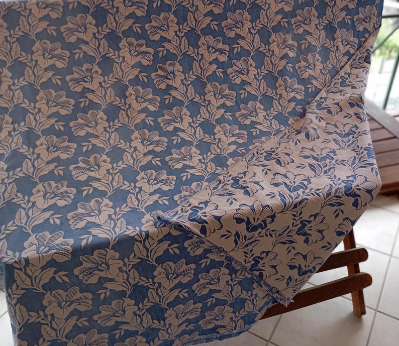 May include: A blue and white floral patterned fabric with a repeating design of stylized flowers. The fabric is folded in half and draped over a wooden chair.