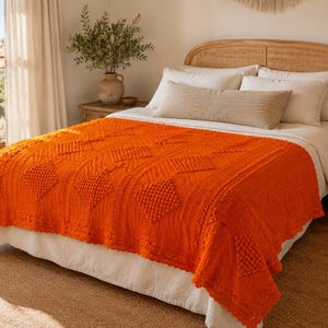May include: A vibrant orange knitted throw blanket featuring a textured diamond pattern with raised bobble details. The blanket is draped over a bed with white bedding, showcasing a detailed cable knit border and a soft, woven construction.