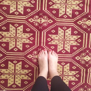 May include: A pair of bare feet with red nail polish stand on a red and gold patterned rug. The rug has a geometric design with repeating stars.