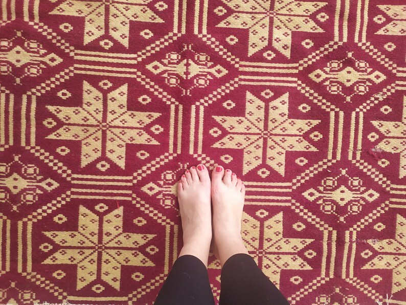 May include: A pair of bare feet with red nail polish stand on a red and gold patterned rug. The rug has a geometric design with repeating stars.