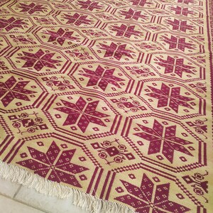 May include: A beige and burgundy patterned rug with a fringe border. The rug features a repeating geometric design with stars and other shapes.