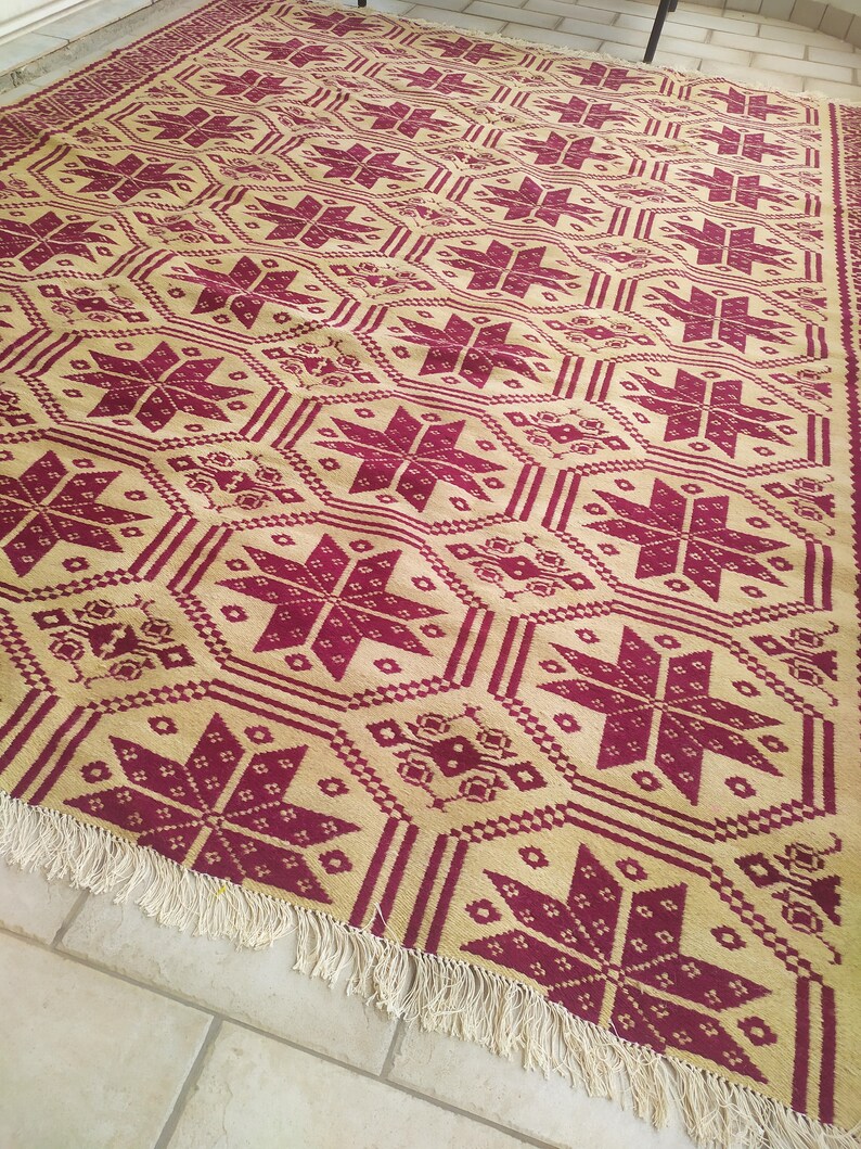 May include: A beige and burgundy patterned rug with a fringe border. The rug features a repeating geometric design with stars and other shapes.