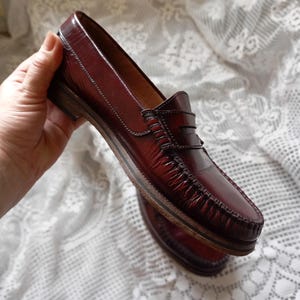 May include: Burgundy leather loafers with a classic penny loafer design. The shoes feature a stitched vamp and a low heel. The leather has a glossy finish, and the soles are made of a light-colored material.