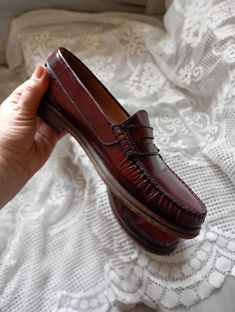 May include: Burgundy leather loafers with a classic penny loafer design. The shoes feature a stitched vamp and a low heel. The leather has a glossy finish, and the soles are made of a light-colored material.