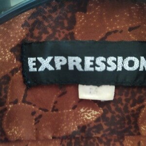 Puede incluir: Una etiqueta negra con costuras blancas que dice "EXPRESSIONS&reg;" sobre una tela de estampado marr&oacute;n.