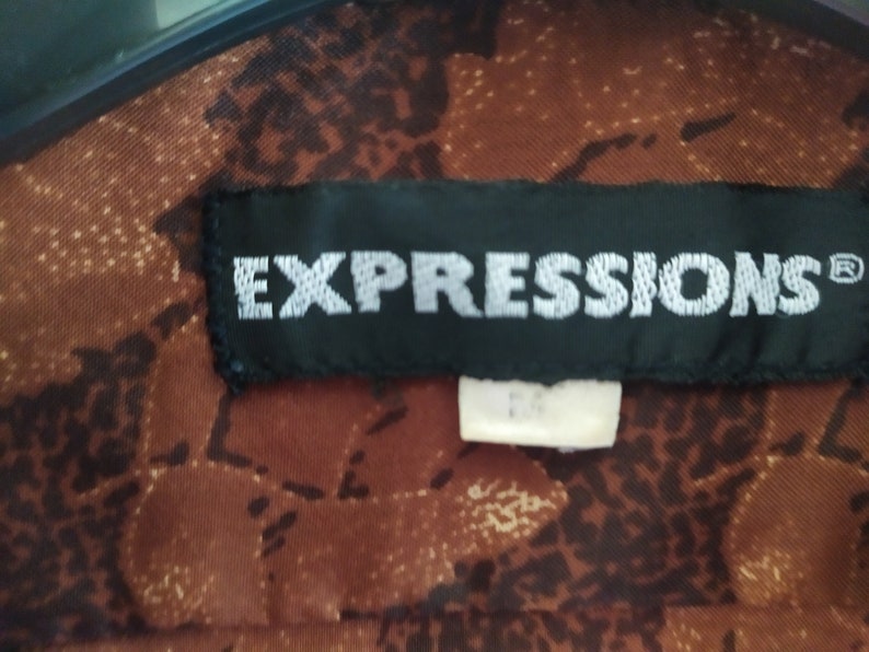 Puede incluir: Una etiqueta negra con costuras blancas que dice "EXPRESSIONS&reg;" sobre una tela de estampado marr&oacute;n.
