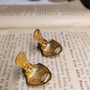 May include: A pair of gold-tone clip-on earrings with a shield-shaped design. The earrings have a textured surface and a detailed design.
