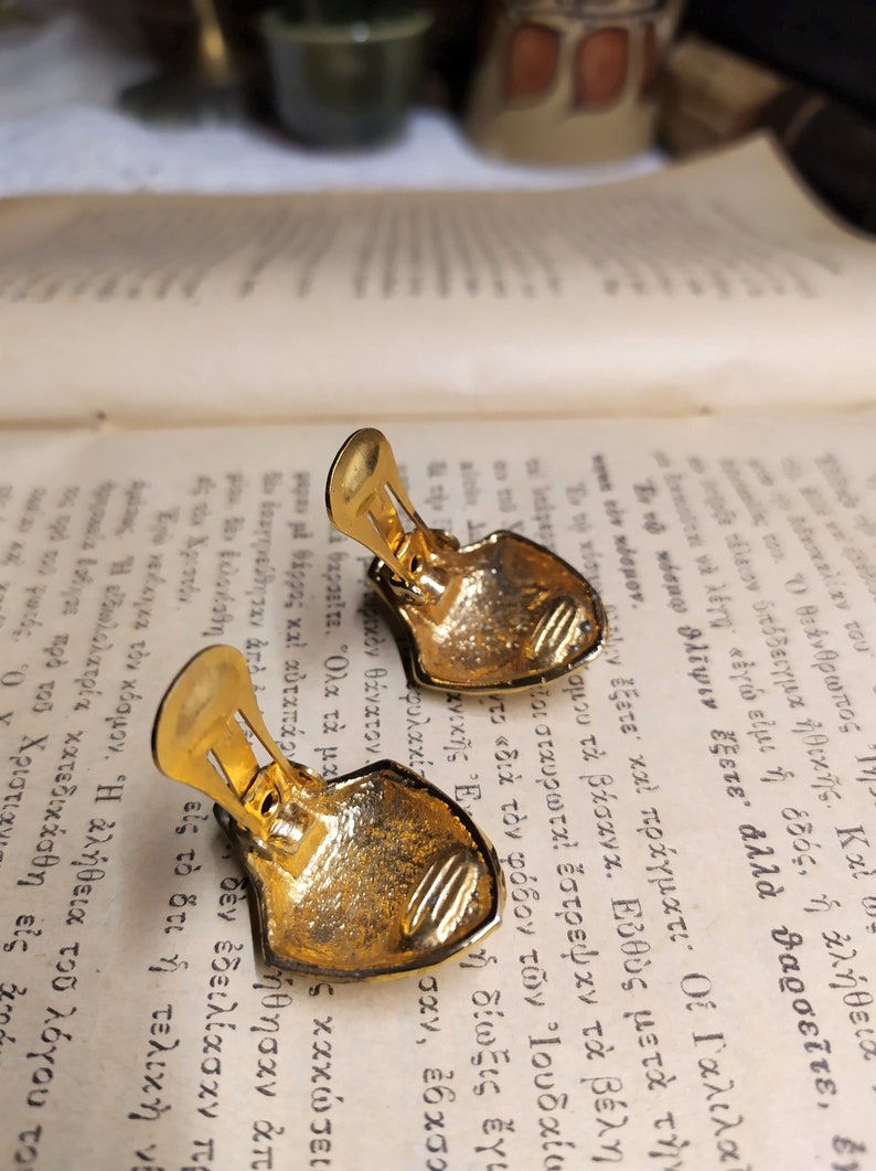 May include: A pair of gold-tone clip-on earrings with a shield-shaped design. The earrings have a textured surface and a detailed design.