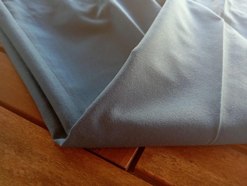May include: A folded piece of smooth, matte, slate-grey fabric with a visible stitched hem along the edge. The material has a uniform, solid colour and a soft texture, showing clean lines and folds where the fabric is layered over itself.