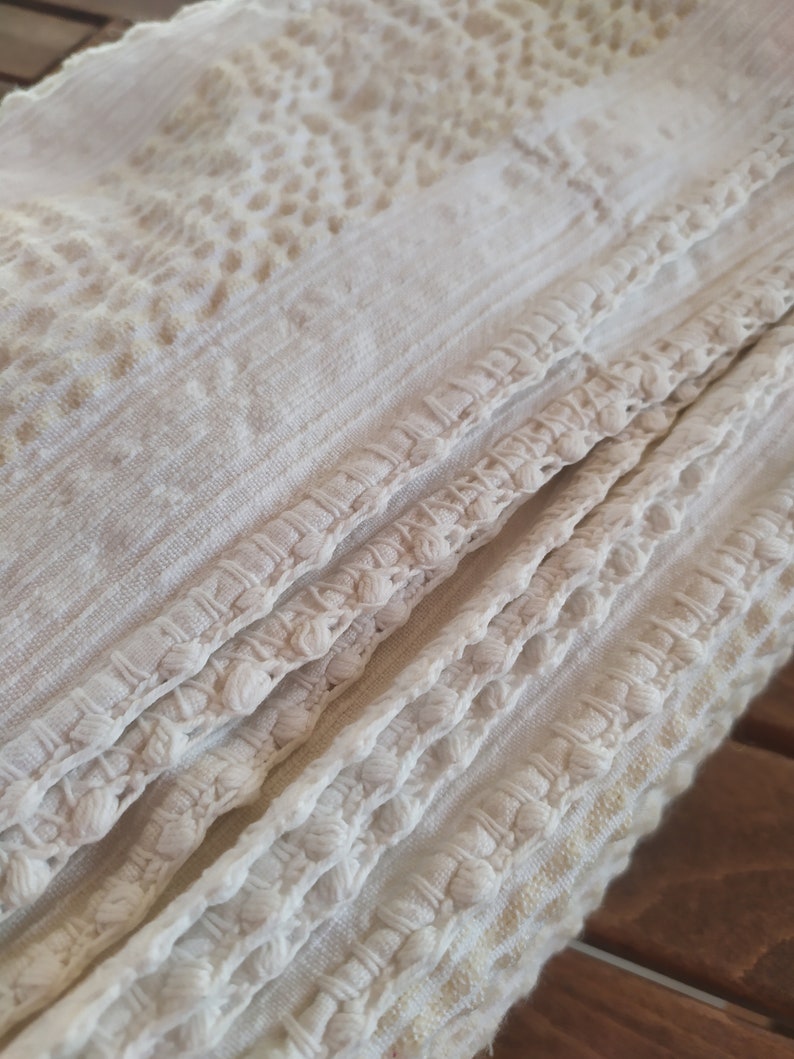 May include: White crocheted fabric with a textured pattern and a fringe border. The fabric is folded in thirds, showing the intricate detail of the crocheted design.