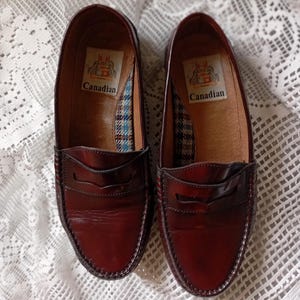 May include: A pair of burgundy leather penny loafers. The shoes have a classic design with a stitched vamp and a decorative strap across the top. The interior features a plaid pattern and the word "Canadian".