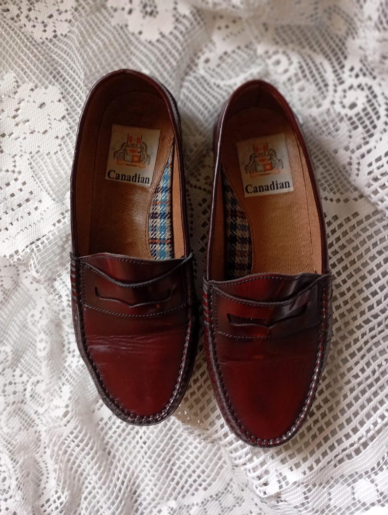 May include: A pair of burgundy leather penny loafers. The shoes have a classic design with a stitched vamp and a decorative strap across the top. The interior features a plaid pattern and the word "Canadian".