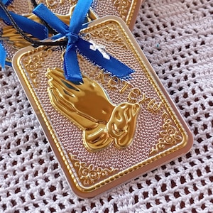 Op de afbeelding: Een rechthoekig zilveren en gouden frame met een reliëfdesign en een gouden afbeelding van gevouwen handen. Het frame heeft een blauw lint eromheen gebonden met een zilveren hanger.
