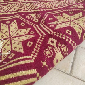 May include: A close-up of a red and gold patterned rug with a geometric design. The rug is folded in half and the edges are visible.