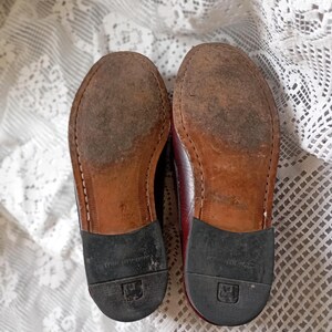 May include: A pair of worn burgundy leather shoes, soles facing upwards. The soles are brown with visible wear and stitching. Black rubber heels are attached. The shoes are on a white lace tablecloth.