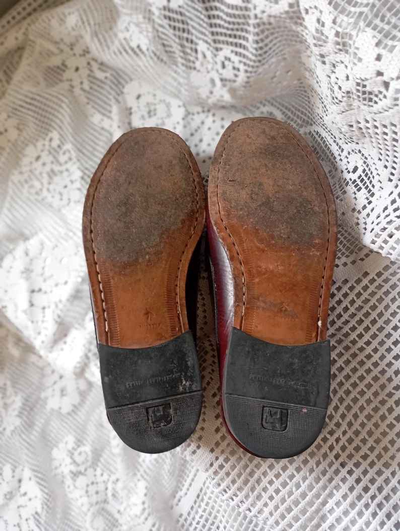 May include: A pair of worn burgundy leather shoes, soles facing upwards. The soles are brown with visible wear and stitching. Black rubber heels are attached. The shoes are on a white lace tablecloth.
