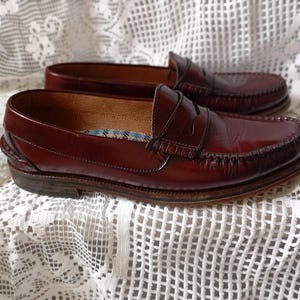 May include: A pair of burgundy leather penny loafers. The shoes feature a classic design with a stitched vamp, a leather sole, and a decorative strap. The interior lining has a blue and white plaid pattern.