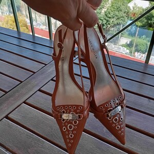 May include: A pair of brown leather heeled shoes with a pointed toe and a silver buckle. The shoes have a decorative pattern of small silver circles on the toe and heel. The shoes are labeled "Karen Millen London".