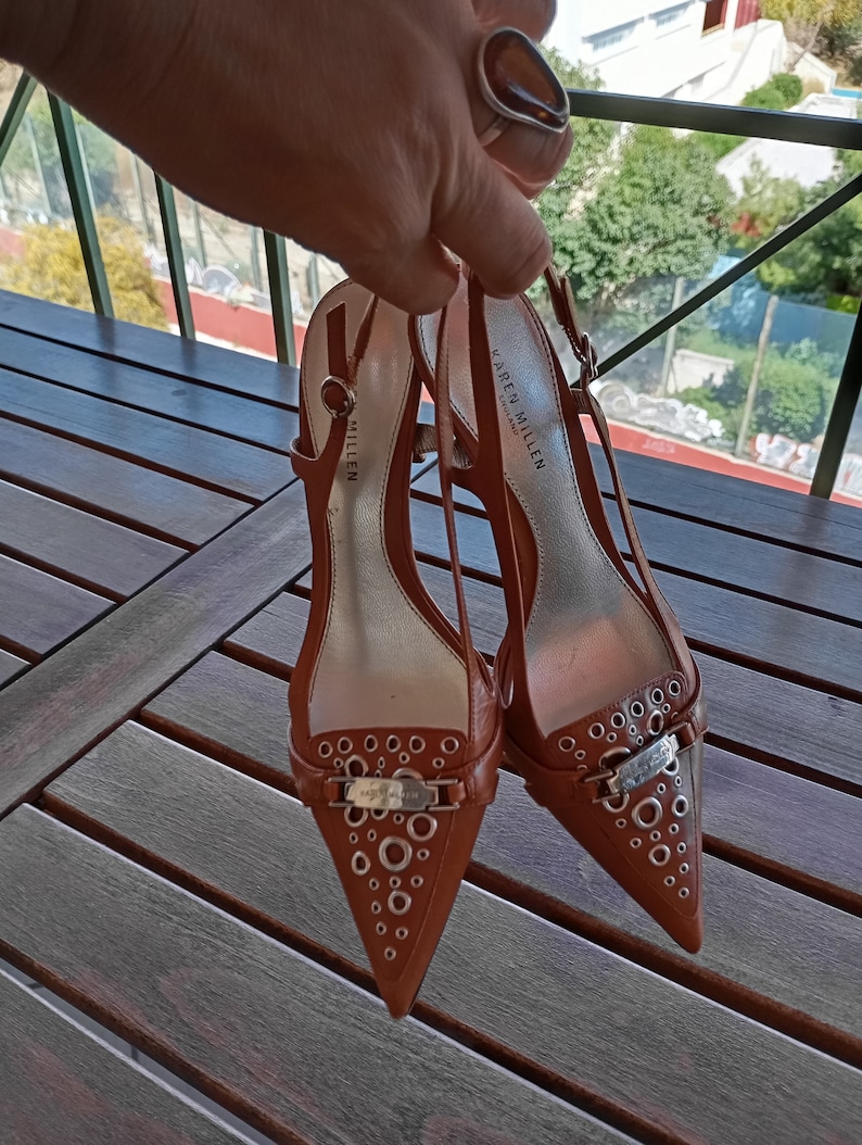 May include: A pair of brown leather heeled shoes with a pointed toe and a silver buckle. The shoes have a decorative pattern of small silver circles on the toe and heel. The shoes are labeled "Karen Millen London".
