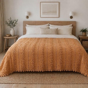 Op de afbeelding: Een slaapkamer met een bed bedekt met een getextureerde, oranje deken. Het bed heeft wit beddengoed en een houten hoofdbord. De kamer is ingericht in neutrale tinten, met een jute vloerkleed en houten meubels, wat een gezellige sfeer creëert.