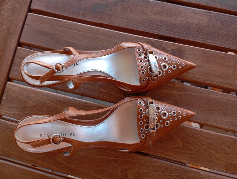 May include: A pair of brown leather heeled shoes with silver buckles and eyelets. The shoes have a pointed toe and an ankle strap. The brand name "Karen Millen" is visible on the sole of one shoe.