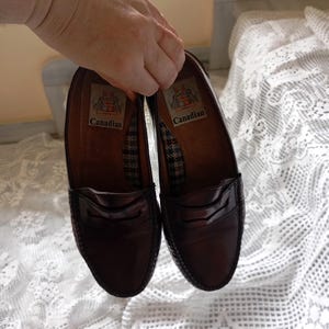 May include: A pair of dark brown leather penny loafers. The shoes have a glossy finish and a classic design with a strap across the vamp. The insoles are brown with a patterned fabric and the word "Canadian".