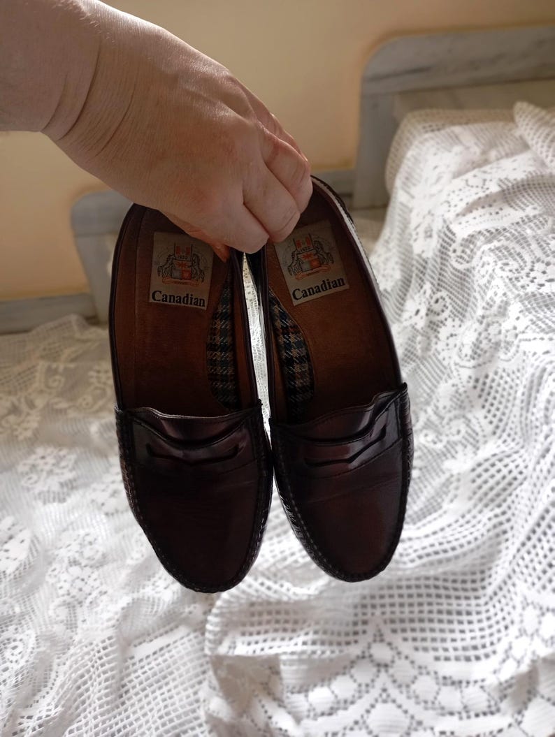 May include: A pair of dark brown leather penny loafers. The shoes have a glossy finish and a classic design with a strap across the vamp. The insoles are brown with a patterned fabric and the word "Canadian".