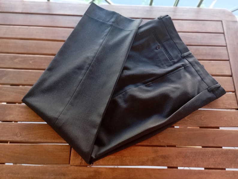 May include: A pair of dark charcoal grey dress trousers featuring a structured waistband with belt loops, a single visible button closure, and a clean, pressed front crease. The fabric has a smooth, matte finish with tailored seams.