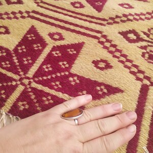 May include: A close-up of a hand wearing a silver ring with an amber stone. The hand is resting on a woven rug with a geometric pattern in shades of red and beige.
