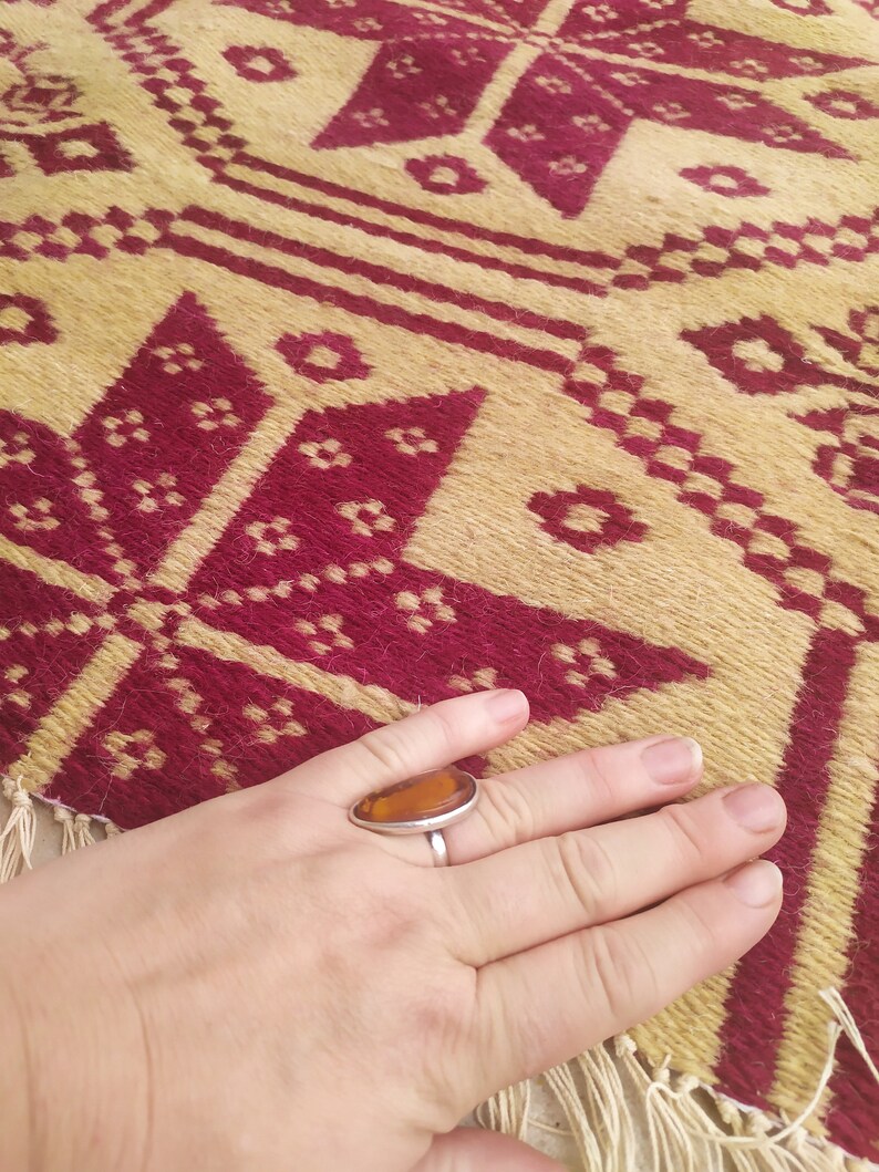 May include: A close-up of a hand wearing a silver ring with an amber stone. The hand is resting on a woven rug with a geometric pattern in shades of red and beige.