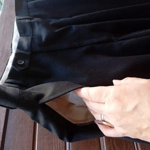 May include: Black trousers featuring a side pocket with a light beige lining, a waistband with a buttoned tab closure, and a pleated front design. The fabric has a smooth texture, and the garment includes a visible interior pocket construction.