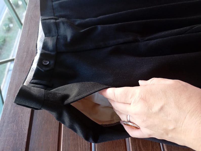 May include: Black trousers featuring a side pocket with a light beige lining, a waistband with a buttoned tab closure, and a pleated front design. The fabric has a smooth texture, and the garment includes a visible interior pocket construction.