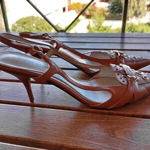 May include: A pair of brown leather heeled shoes with a buckle and eyelet detail. The shoes are on a wooden surface.