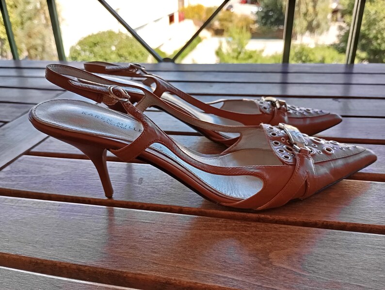 May include: A pair of brown leather heeled shoes with a buckle and eyelet detail. The shoes are on a wooden surface.