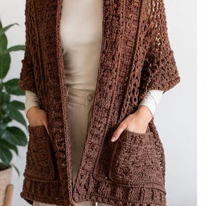 May include: A dark brown crochet cardigan featuring an open front, short sleeves, and two large front pockets. The garment is crafted with a textured, repeating stitch pattern throughout, creating a structured yet flexible look with a wide collar.