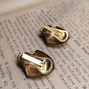 May include: A pair of gold-tone clip-on earrings with a textured, ornate design. The earrings are shaped like a shield with a rounded top.