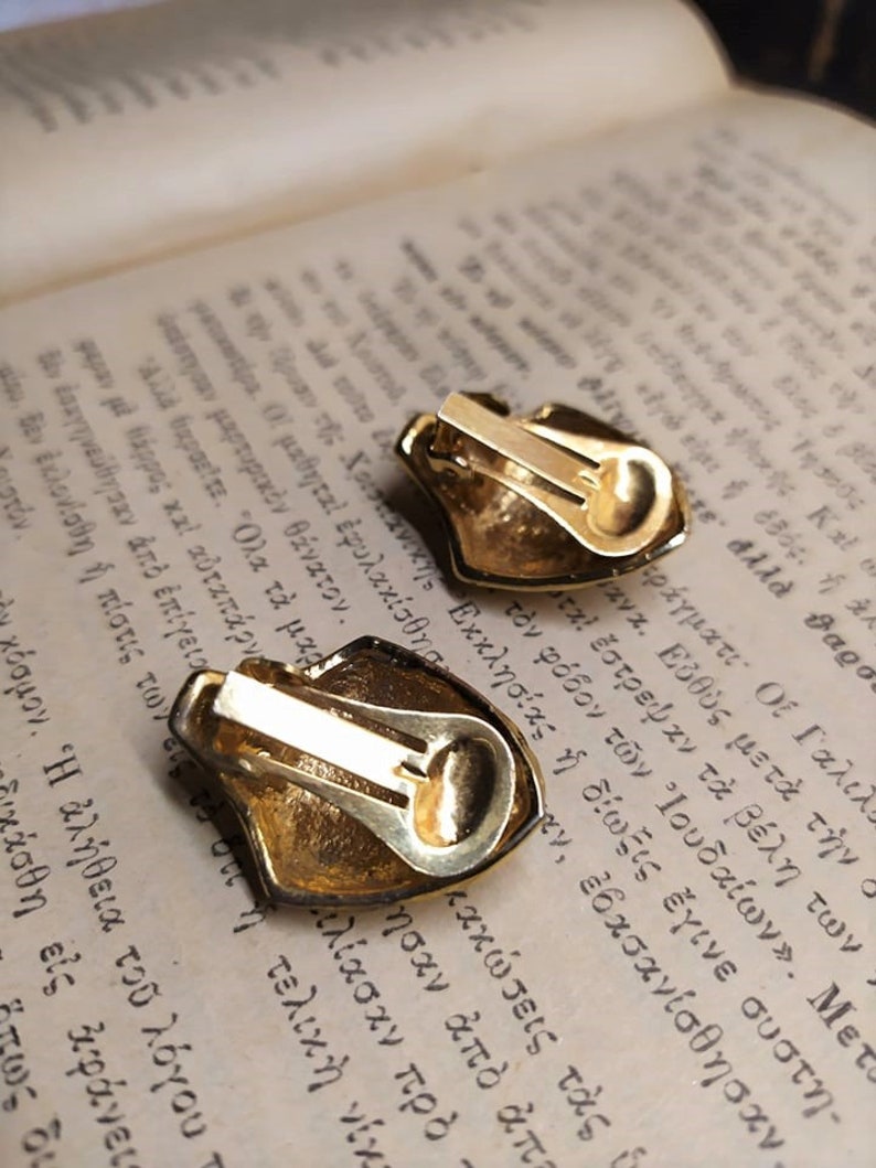 May include: A pair of gold-tone clip-on earrings with a textured, ornate design. The earrings are shaped like a shield with a rounded top.