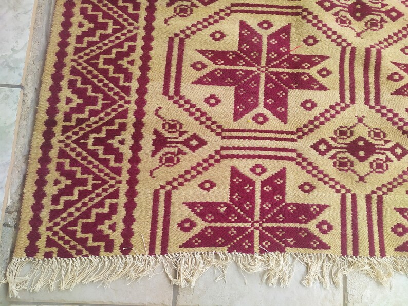 May include: A beige and burgundy rug with a geometric pattern. The rug has a fringed border.