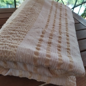 May include: A white and beige striped towel with a textured pattern. The towel is folded in half and is sitting on a wooden surface.