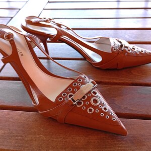 May include: A pair of brown leather heeled shoes with a pointed toe and a silver buckle. The shoes have a cut-out design and are decorated with silver eyelets. The brand name "ALIEN" is visible on the insole.