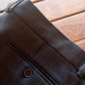 May include: A pair of dark charcoal grey trousers featuring a structured waistband with belt loops and a single welt pocket on the front. The pocket is secured with a small, round, dark-coloured button centred on the horizontal opening.
